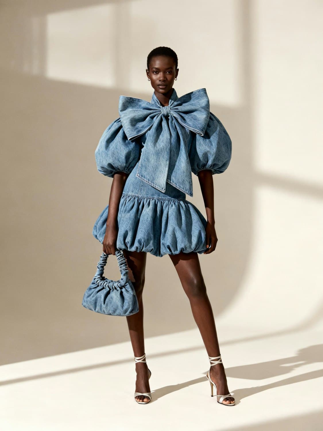 Denim mini dress with puffed sleeves, an oversized bow and a bubble hem, carried with a denim bag