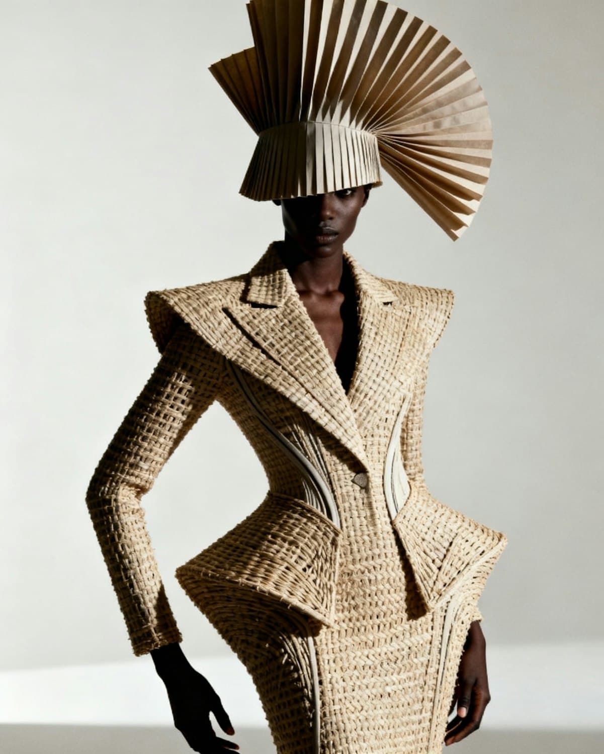 Model in a sculpted woven raffia jacket with a flared peplum and a towering pleated fan headpiece