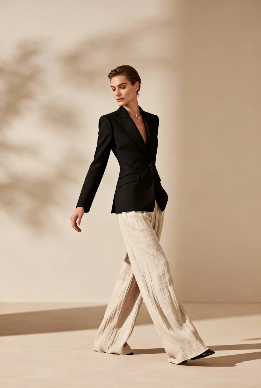 Model walking in a sharp black blazer and wide linen trousers, leaf shadows on the wall behind