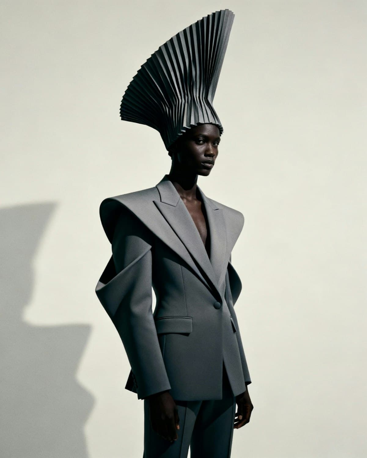 Model in a slate grey suit with sculpted, extended shoulders and a pleated grey fan headpiece