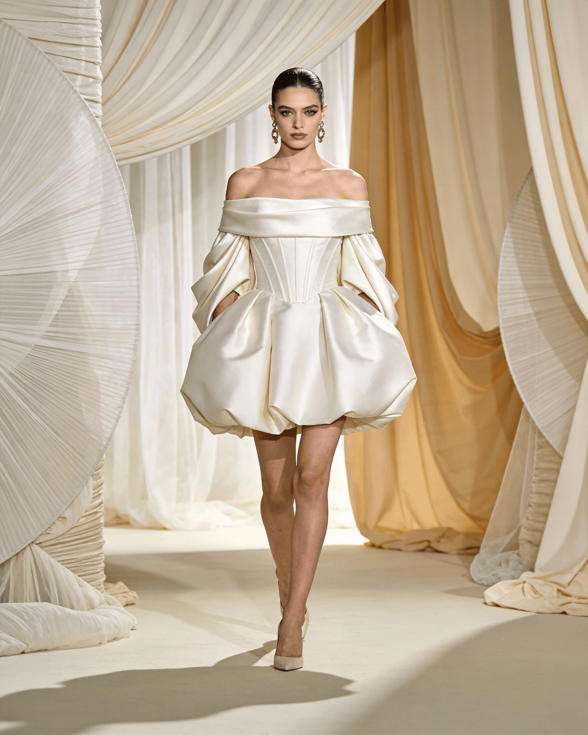 Model in an ivory off-the-shoulder corset dress with a sculpted bubble skirt, walking between cream drapes